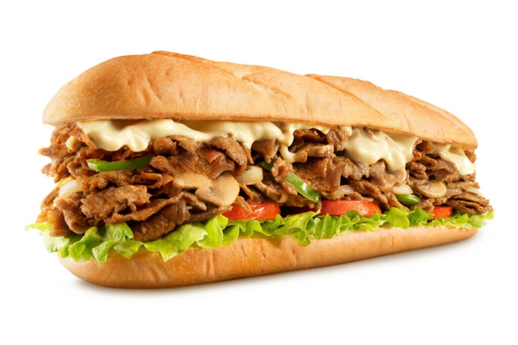 Steak Philly Cheesesteak from Charleys Cheesesteaks against a white background. The Steak Philly Cheesesteak from Charleys Philly cheesesteak restaurants is made with melted provolone cheese, grilled steak, onions, and peppers on a toasted roll, then topped with lettuce, tomato, mayo, and pickle. best philly cheesesteak